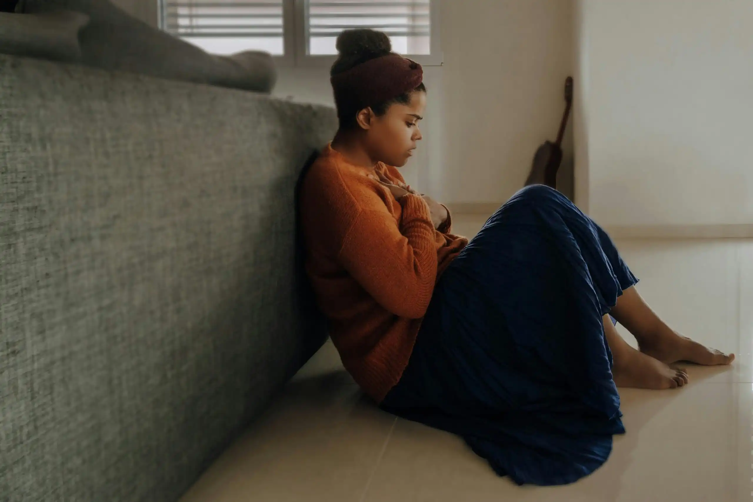 A woman looks anxious while sitting on the floor behind a couch with a blanket over her legs while holding her hands over her chest.