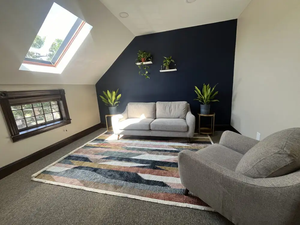 A room with a gray chair, gray sofa, colorful rug, two plants on the floor, and a navy blue wall with plants on two shelves. On one wall is a brown window with wooden details and above that is an angled wall with a skylight letting sunlight in.