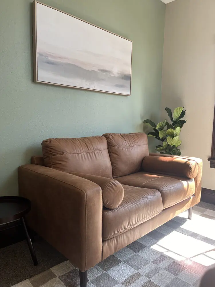 Natural light comes in a room with a light brown leather couch sitting on a muted rug with squares. The wall behind the couch is a light sage green and has an abstract painting with muted and peaceful colors.