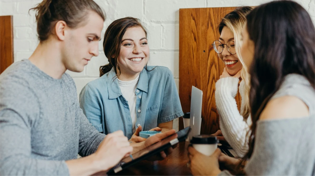 A group of four young people smiling and facing each other. They have coffee cups, a tablet, and a laptop in front of them.