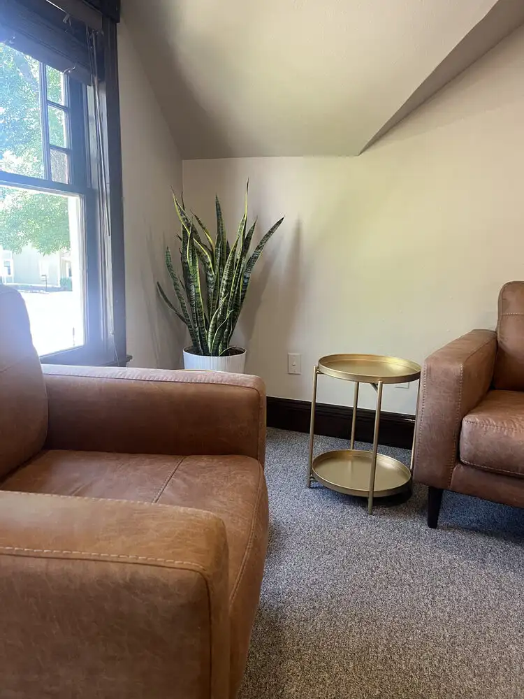 An office with two light brown leather chairs, a gold side table with two levels, and a snake plant in a white pot sits in the corner. A dark wooden window lets natural light in.