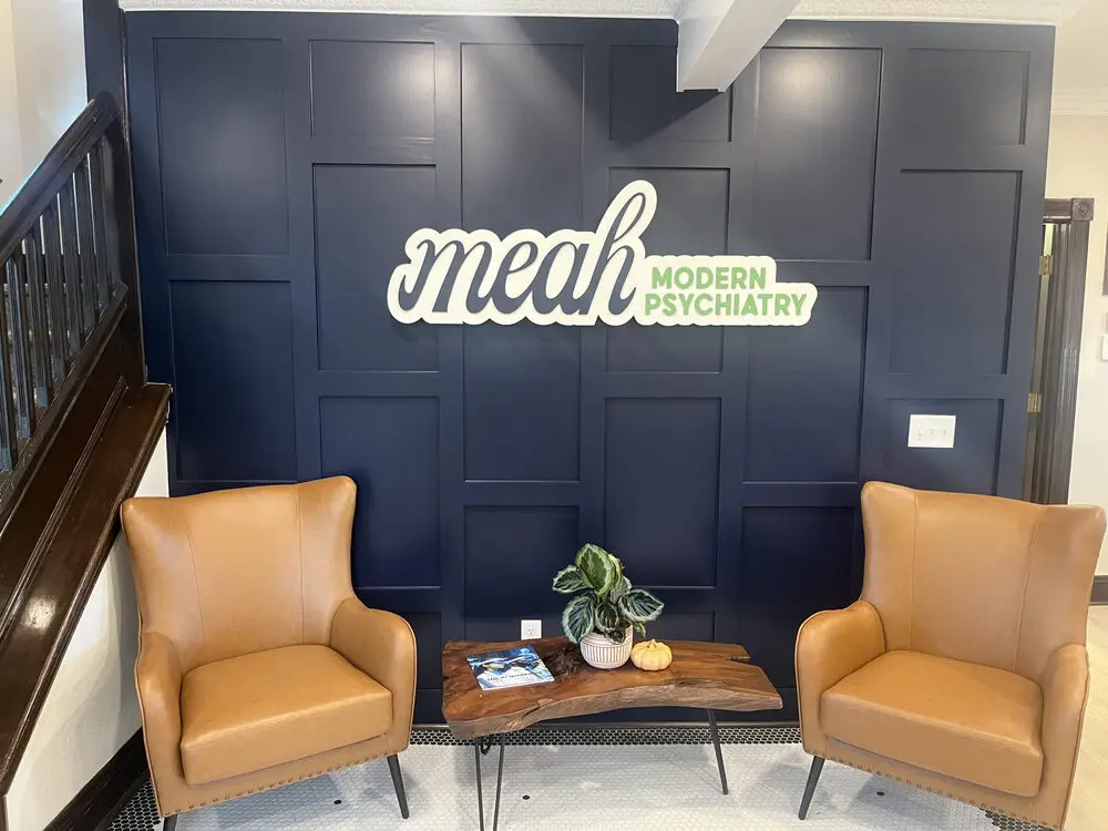 The lobby with two light brown chairs and a wooden table with a plant and magazine sit next to a navy blue wall with a sign reading