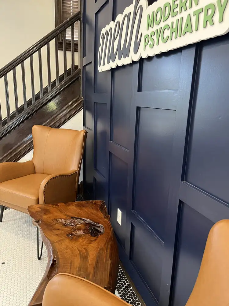 A side shot of the Meah lobby showing a navy blue wall with the Meah Modern Psychiatry sign. In front of the wall sit two light brown chairs and a wooden table sitting on a tile floor. Behind one of the chairs is a dark brown railing to a staircase.