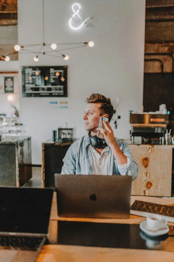 A man sits in a coffee shop in front of a laptop with headphones around his neck and a cell phone up to his hear as he looks off to the side.