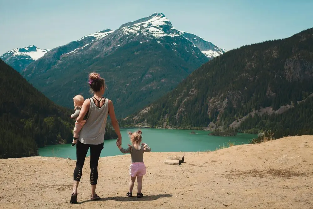 A woman facing away holds a baby and holds the hand of a toddler while they stand on some sand in front of a lake. In the background is a large mountain and some hills covered with trees.